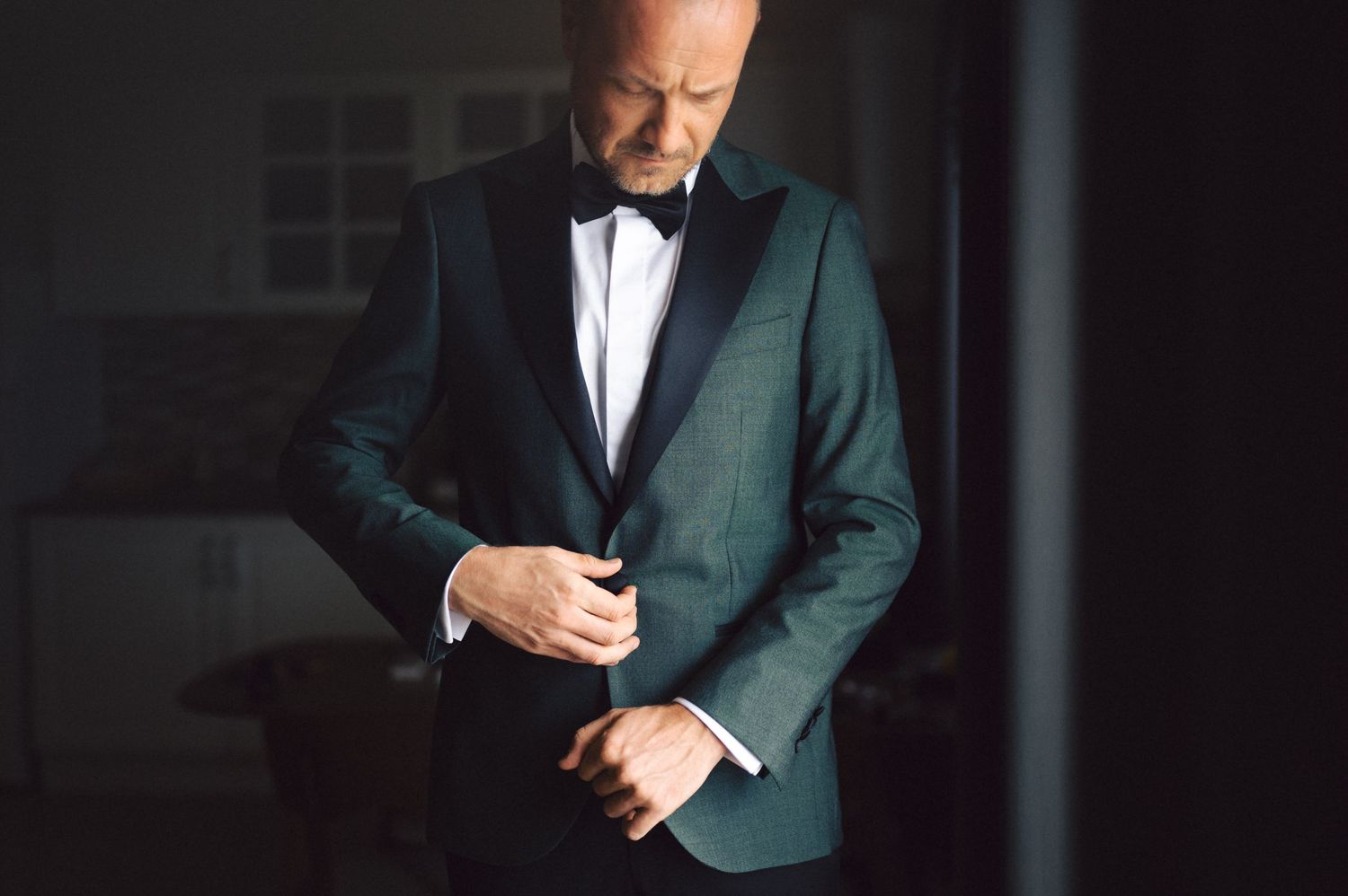 Individual wearing a dark green formal suit adjusts their cufflinks in dramatic lighting.