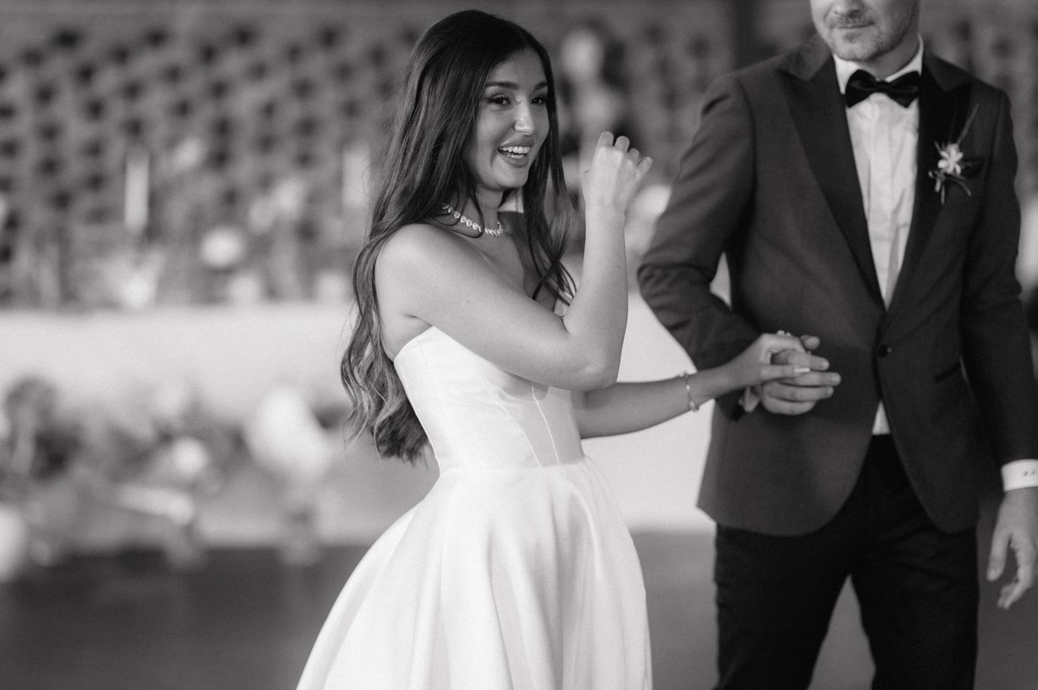 A couple dances together at an event, with the bride wearing a flowing white dress and holding hands with her partner.