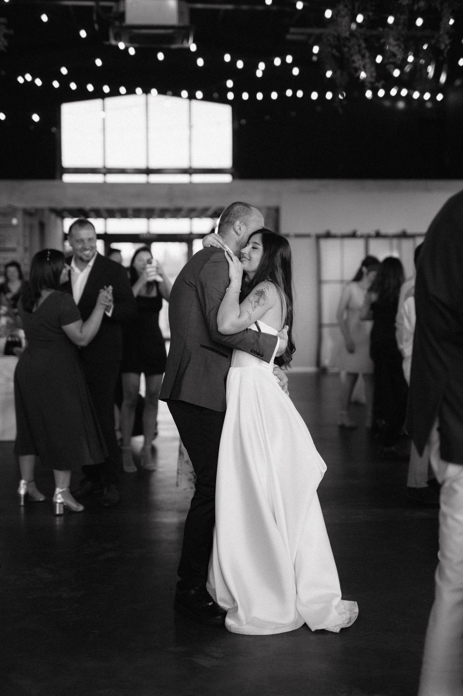 A couple shares a romantic first dance at their wedding reception under twinkling string lights in black and white.