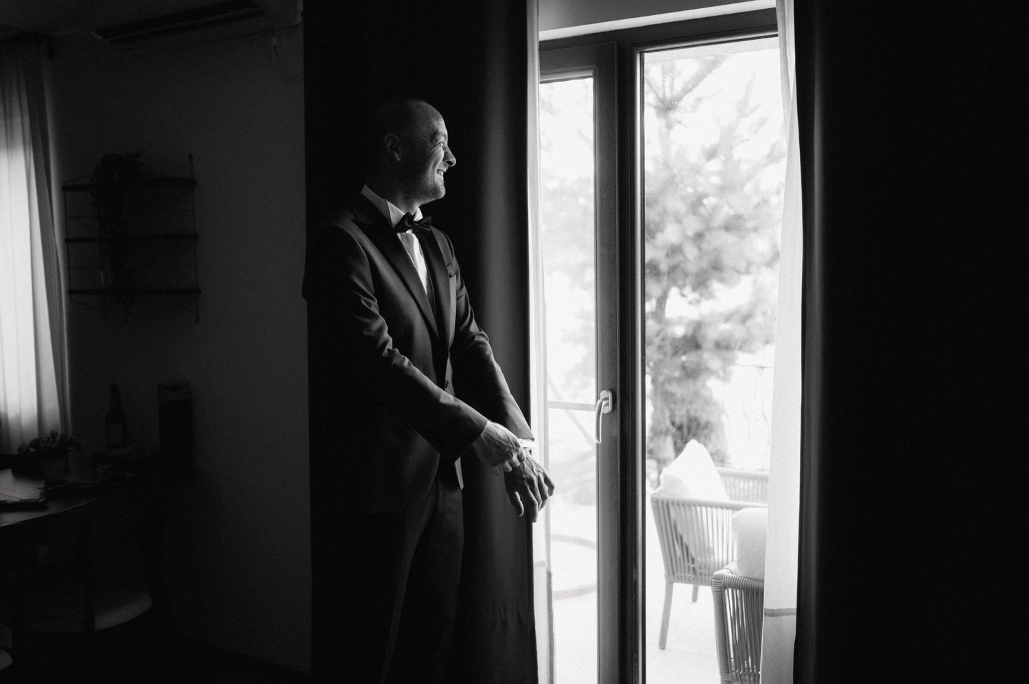 A person in a suit stands in a dark room looking out through a bright glass door in black and white.