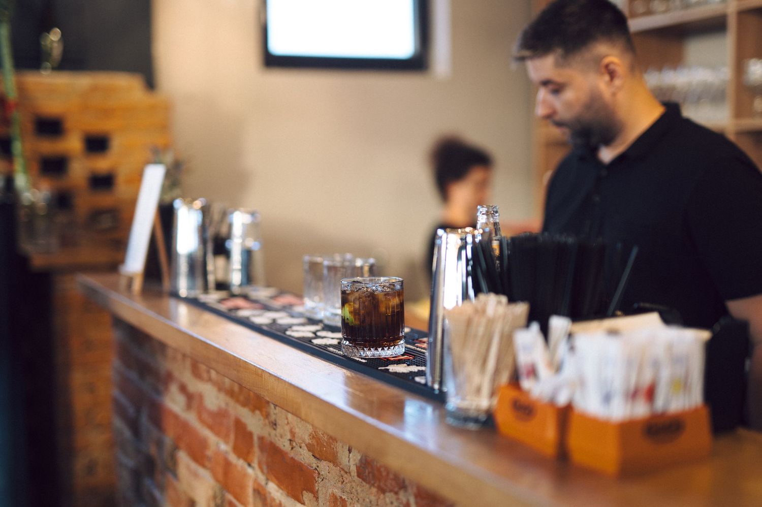 Bartender preparing drinks at a rustic wooden bar counter with glasses and drink equipment visible.