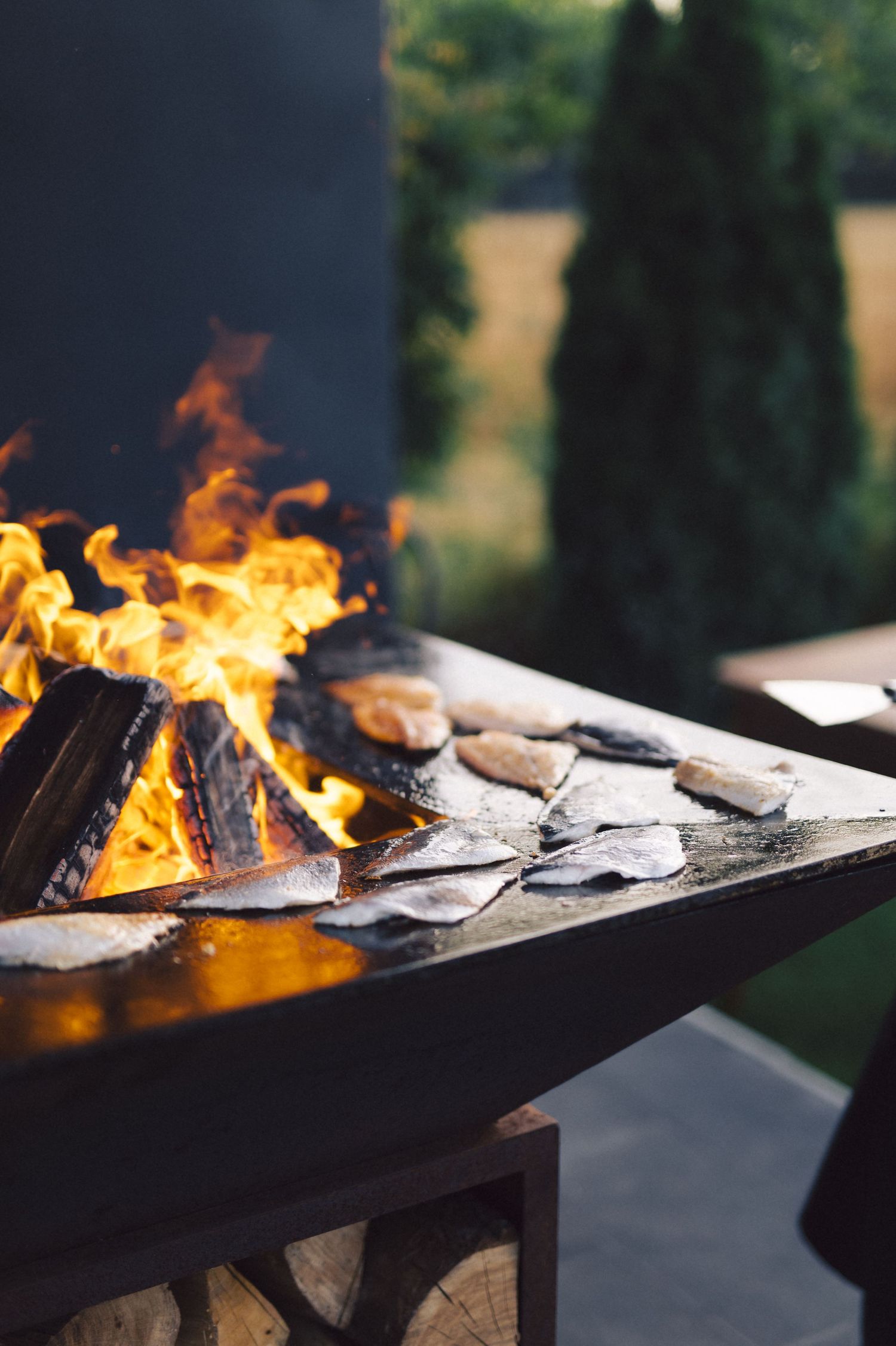 Burgers grilling over open flames on an outdoor barbecue grill with vibrant fire against a blurred green background.