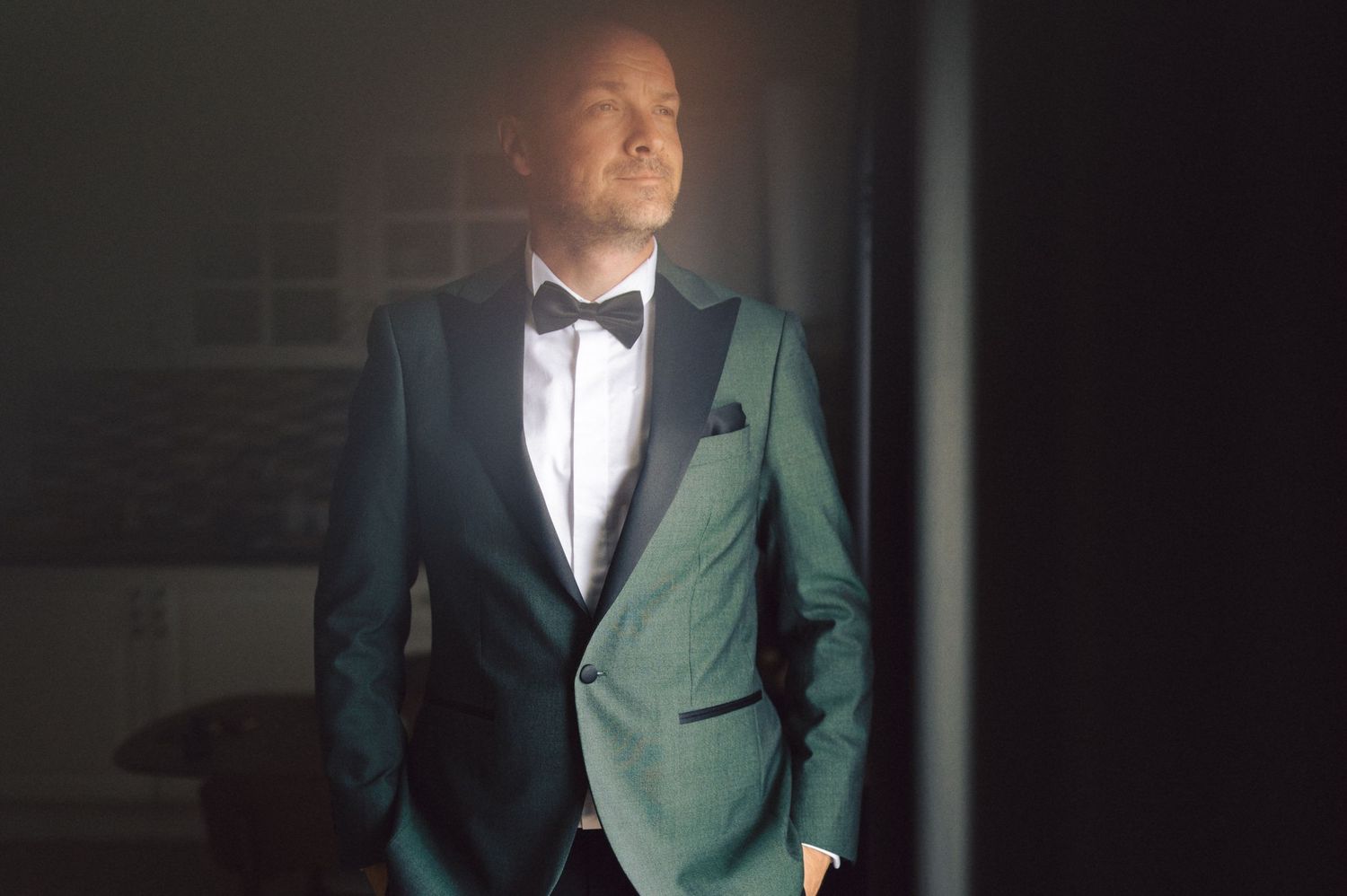 A person stands illuminated in a dark setting wearing a formal teal tuxedo with black lapels and a bow tie.