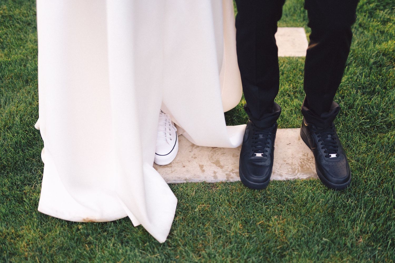 Close-up photo showing elegant bridal gown hem and black dress shoes against green grass.