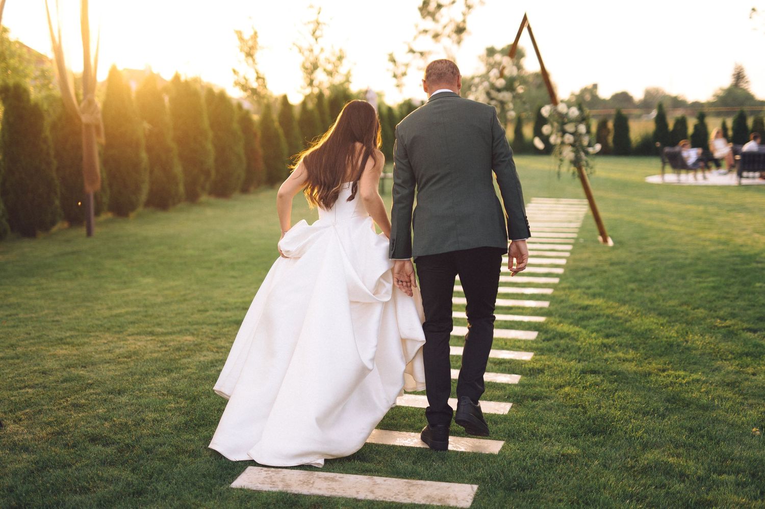 A couple in wedding attire walking down a wooden path at sunset with a scenic outdoor venue in the background.