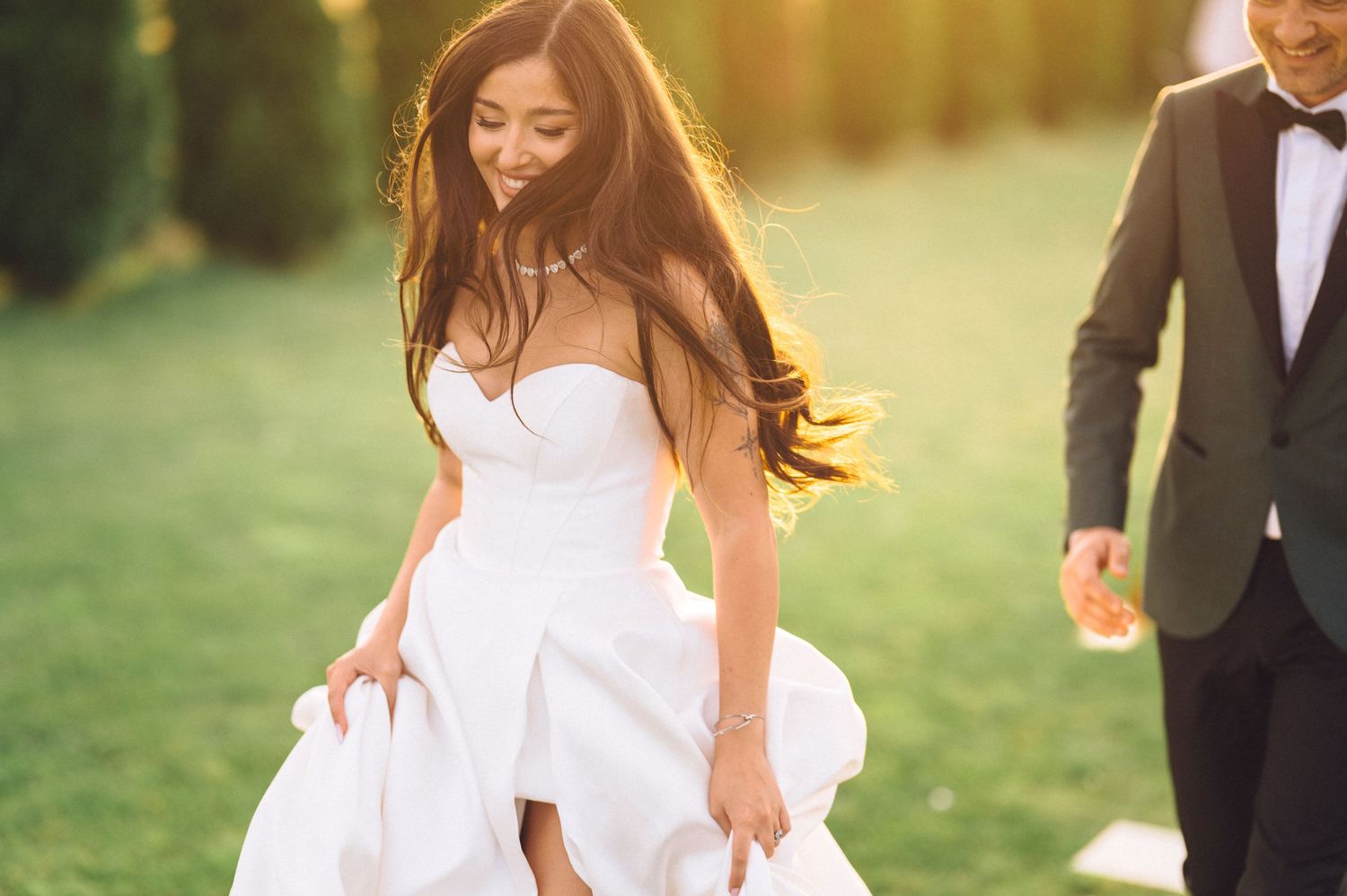 A bride in a flowing white dress walks through a sunlit field while her groom follows behind in a tuxedo.