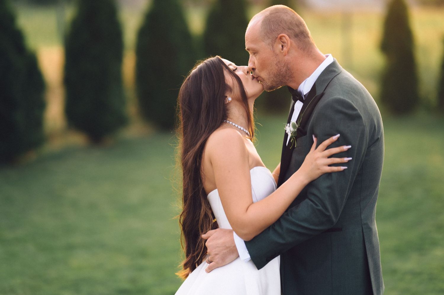 Romantic wedding couple sharing an intimate moment in a garden with blurred cypress trees in the background.