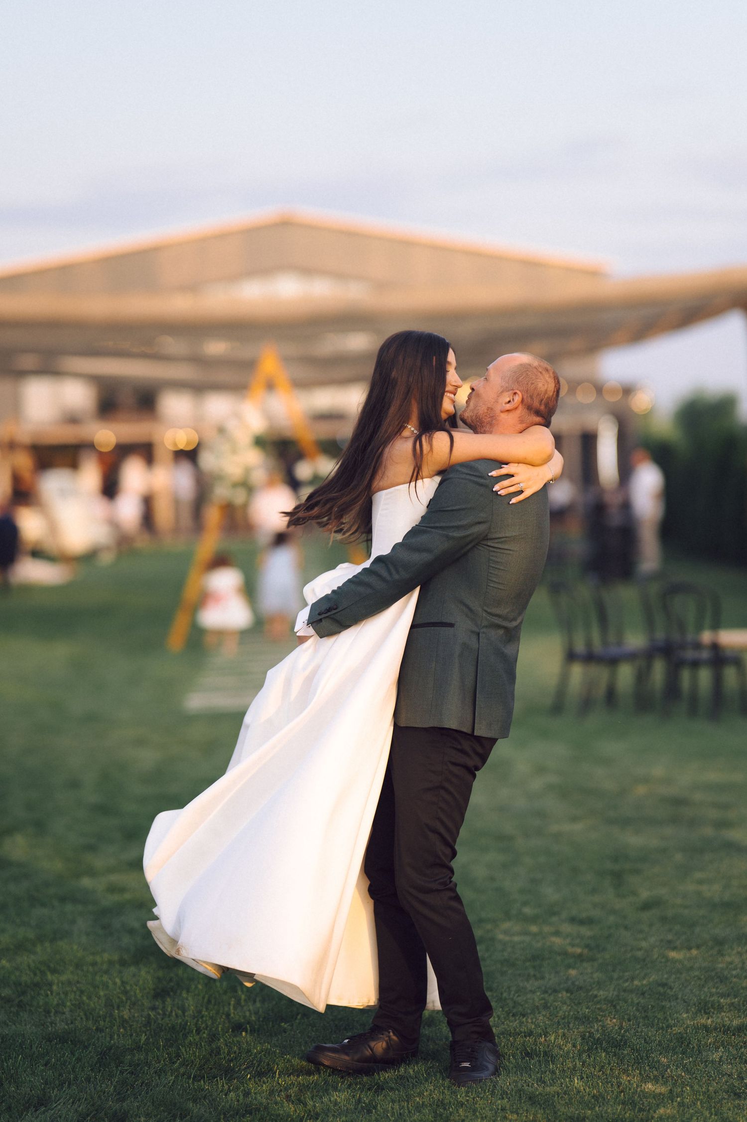 A couple embraces during a wedding celebration on a grassy lawn at sunset.