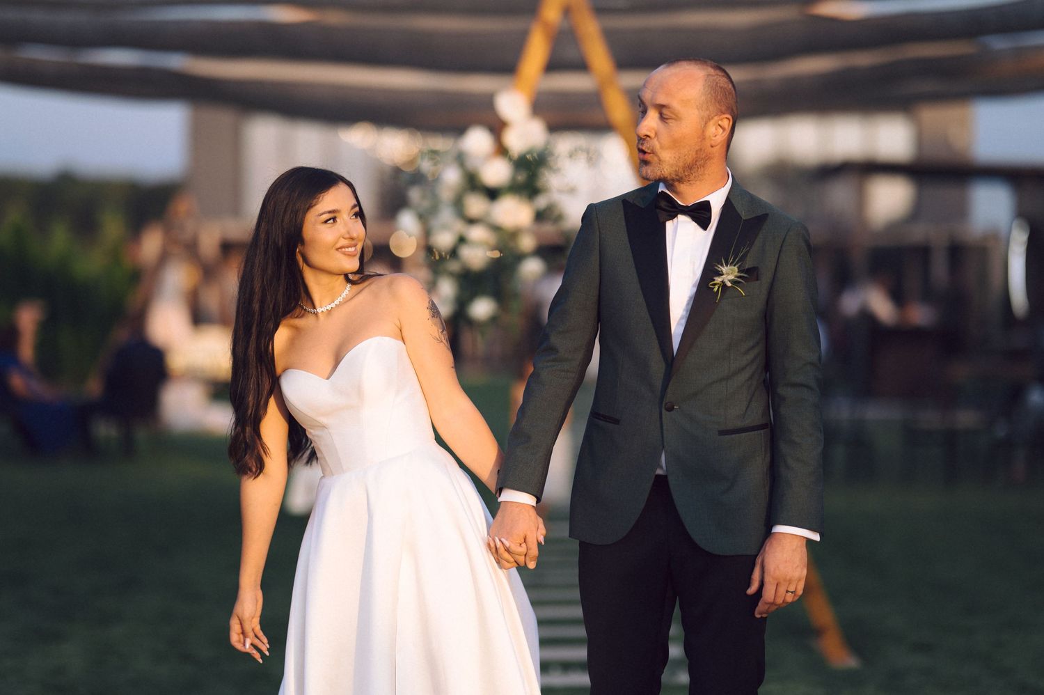 A romantic outdoor wedding scene with the bride in a strapless white gown and groom in classic tuxedo holding hands.