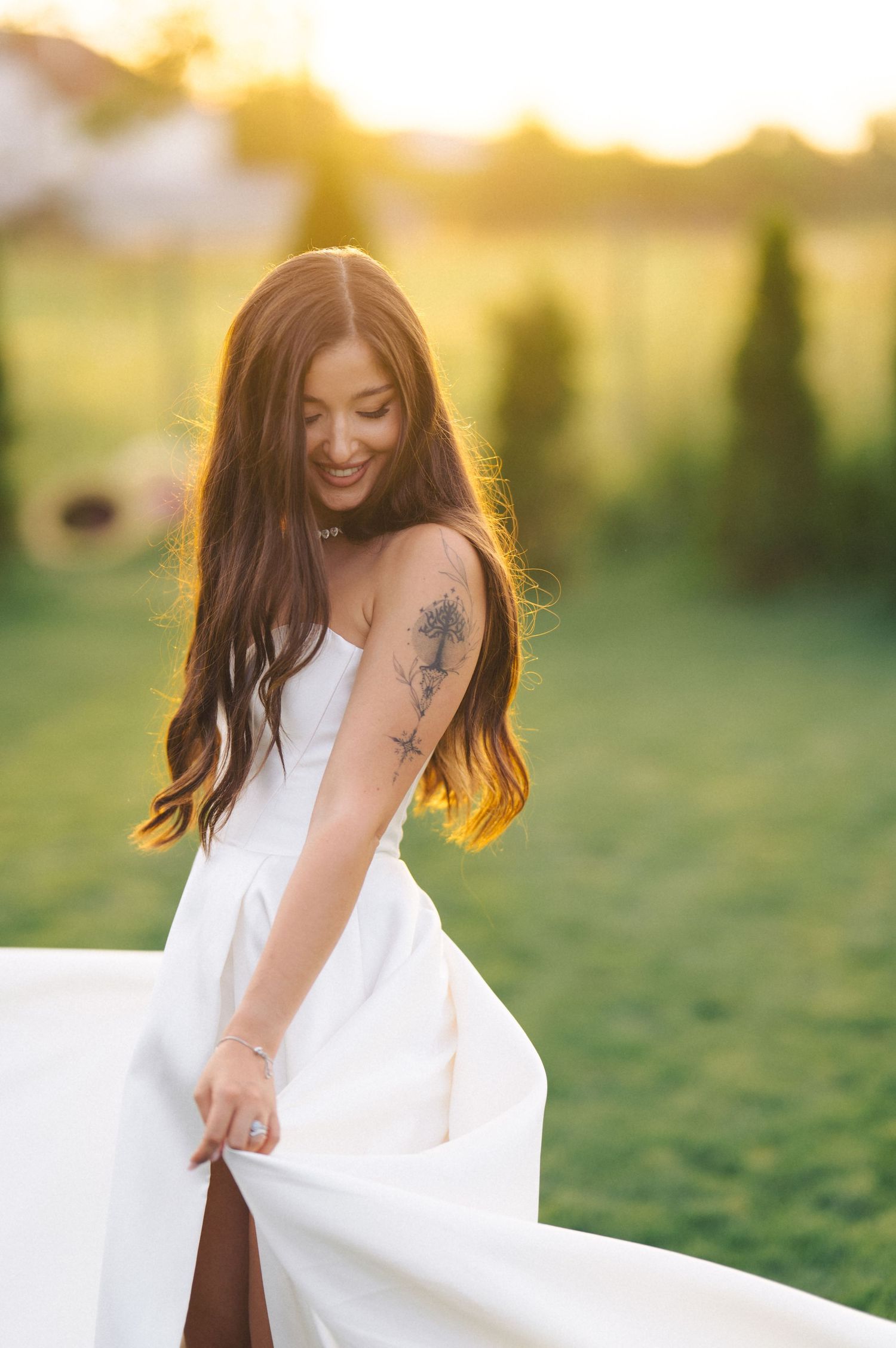 A person in a flowing white dress twirls outdoors at golden hour, showing a tattoo on their arm.