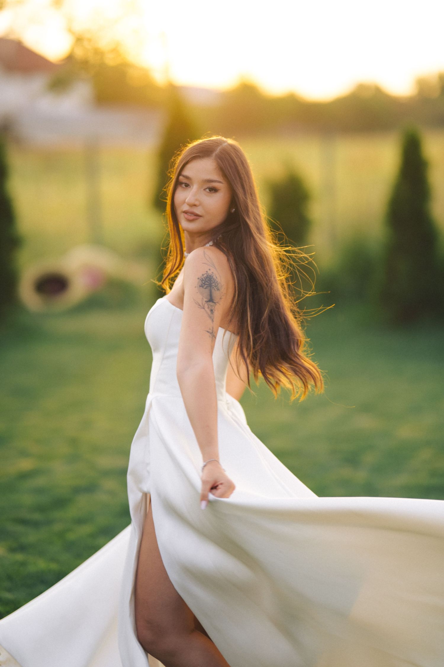 A flowing white dress catches the golden sunlight as long hair flows in the breeze during an outdoor sunset photoshoot.