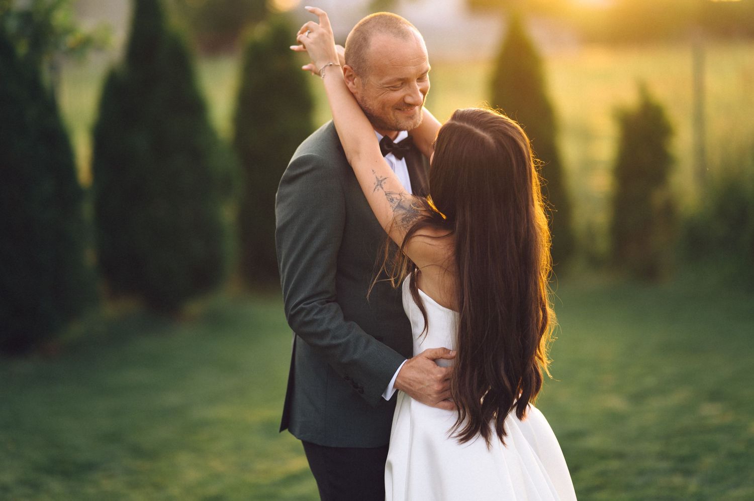 A romantic couple embraces while dancing outdoors at sunset with cypress trees in the background.