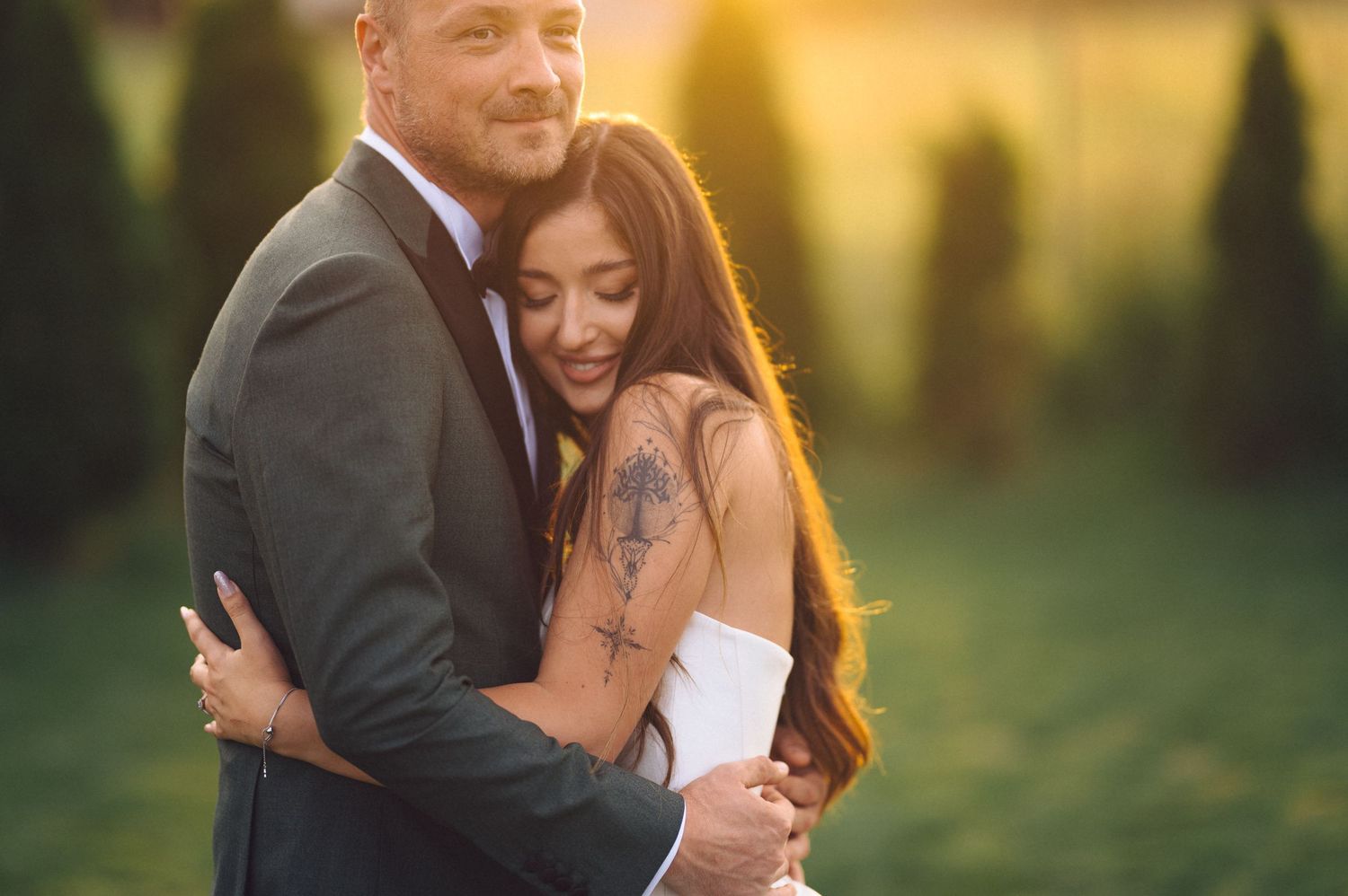 Romantic couple embraces in warm sunset light as bride in white dress shows tattoo on arm during outdoor wedding photoshoot.