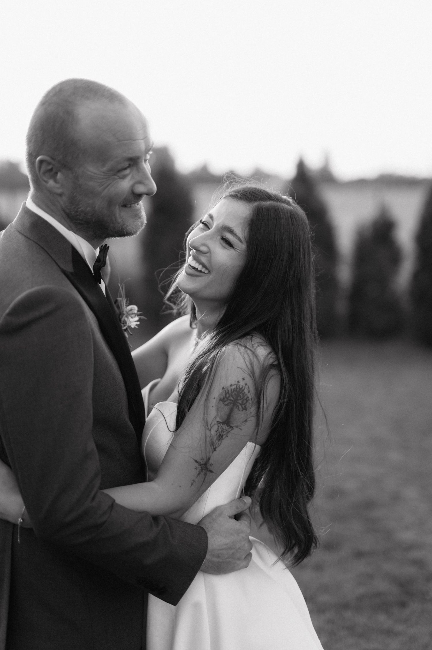 Black and white candid wedding photo showing joyful intimate moment between newlywed couple embracing outdoors.