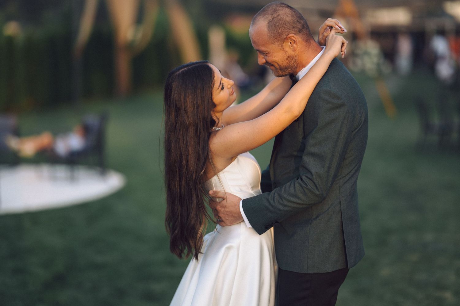 A couple in formal wear share an intimate dance moment outdoors on a green lawn at dusk.