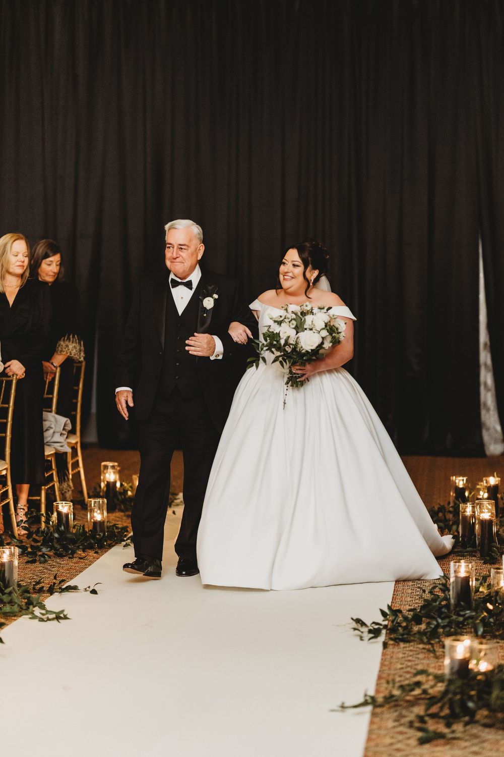 Wedding ceremony with bride and escort walking down candlelit aisle with floral decorations and dark curtained backdrop.