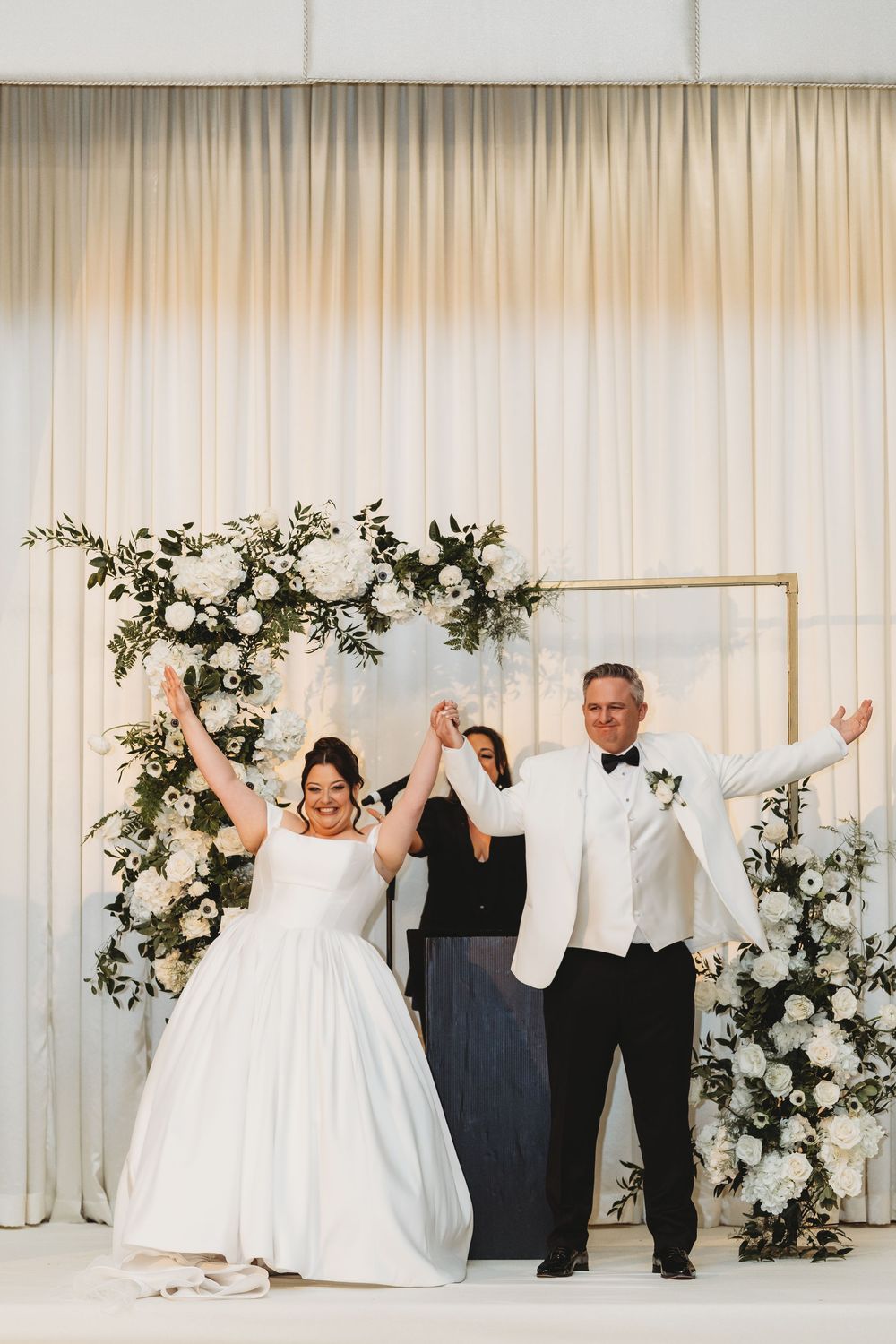 Newlyweds celebrate joyfully with officiant under white floral arch with roses and greenery against draped backdrop.