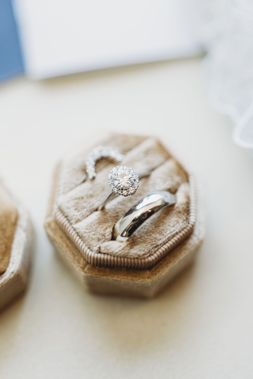 Wedding rings displayed on a beige velvet hexagonal ring box with elegant diamond engagement ring and plain band.