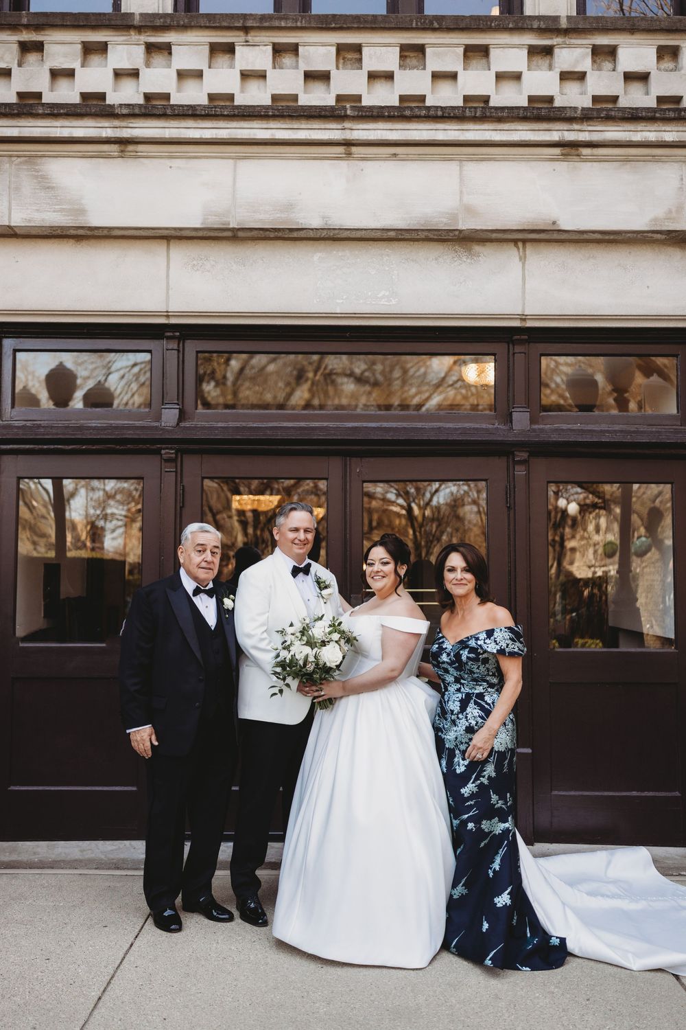 A formal winter wedding group poses outside a classical building with elegant wood and glass doors.