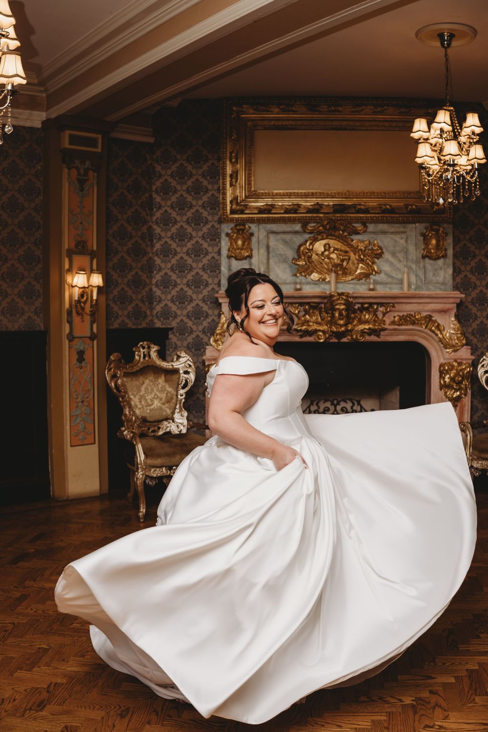 A bride in an elegant off-shoulder white gown twirls in a luxurious room with ornate wallpaper and a decorative fireplace.