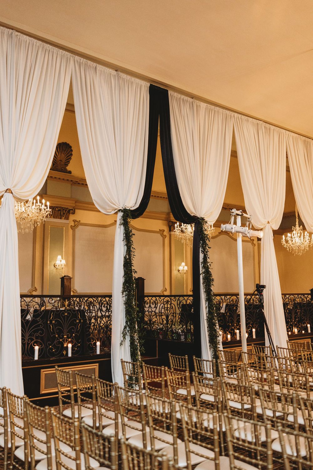 Elegant wedding ceremony setup with white and black drapes, crystal chandeliers, and gold chairs in a formal ballroom.