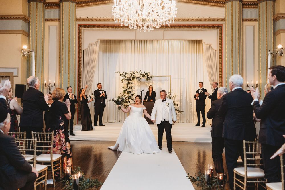 Elegant indoor wedding ceremony in a ballroom with crystal chandelier and white floral arch at the altar.