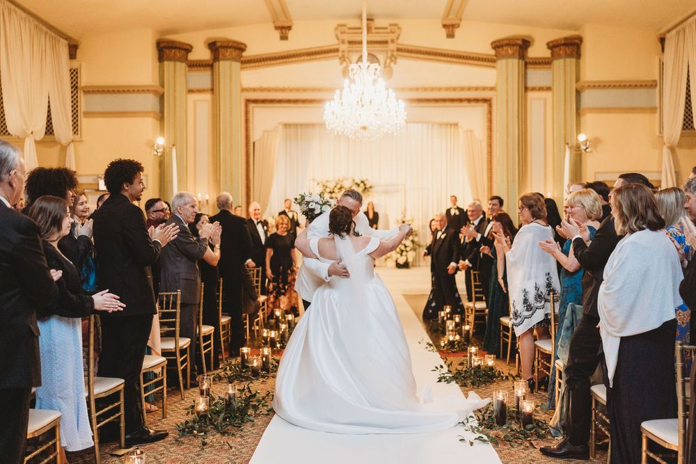 Wedding guests stand during an elegant ceremony in a grand ballroom with crystal chandeliers and gold decor.