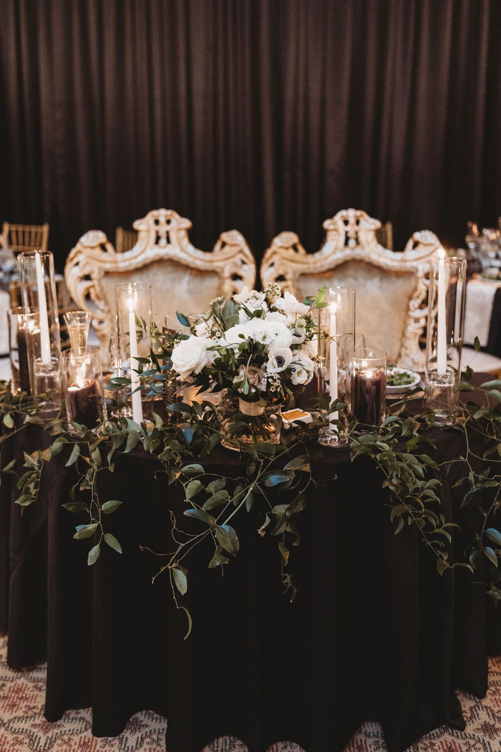 Elegant wedding head table with ornate white chairs, candlelight, and cascading greenery on black tablecloth.