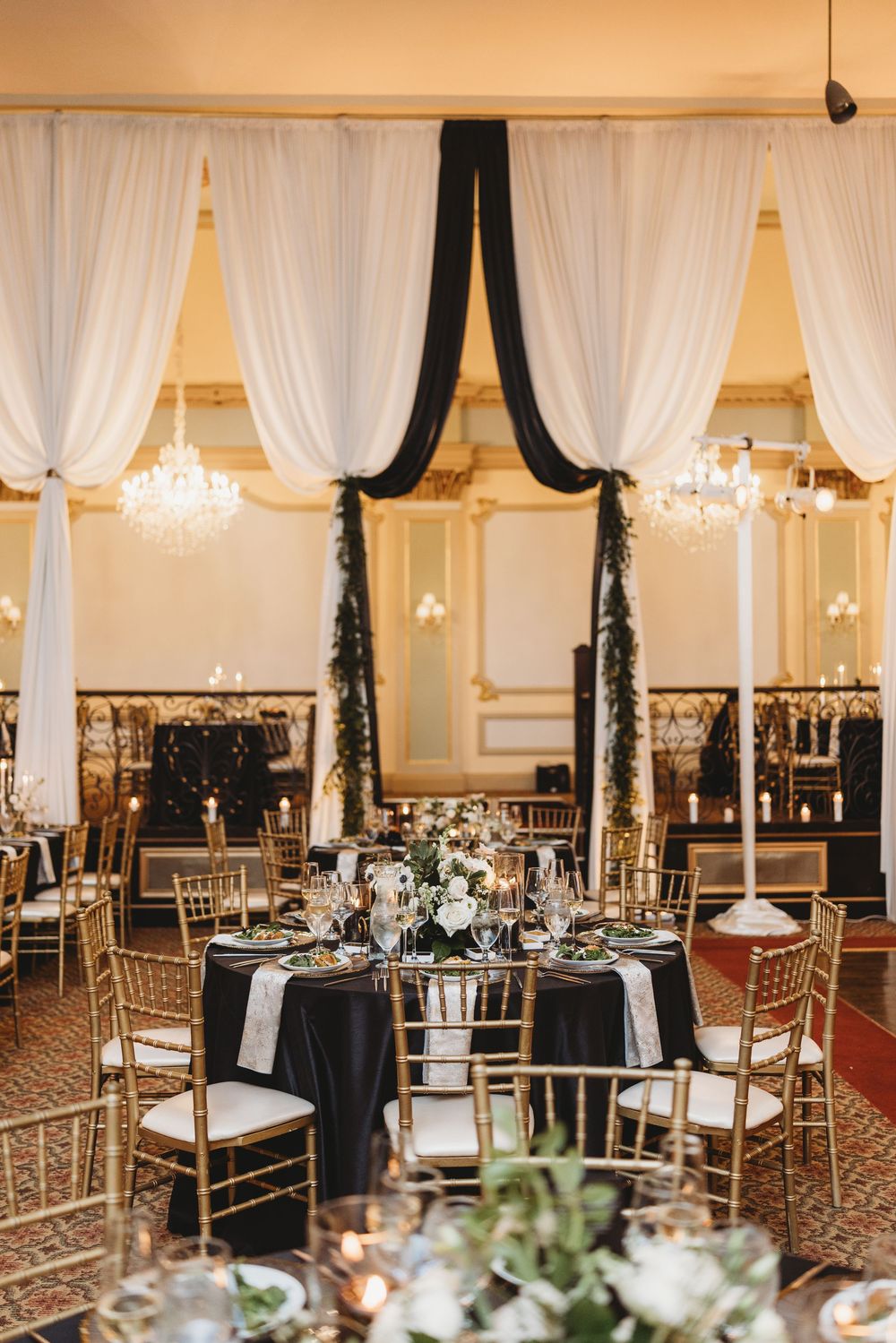 Elegant wedding reception hall with white drapes, crystal chandeliers, gold Chiavari chairs and black tablecloths.