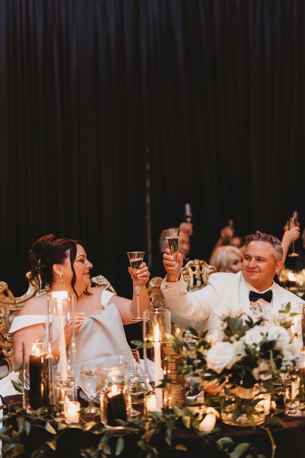 Candlelit wedding reception table with guests seated alongside glowing centerpieces and elegant floral arrangements.