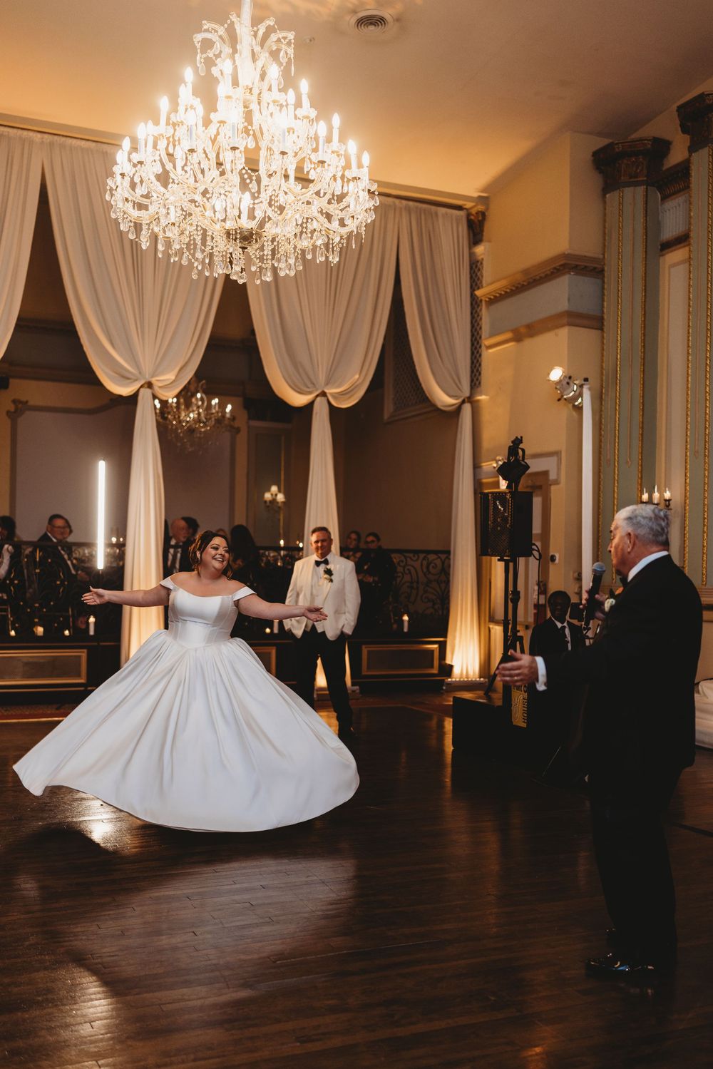A flowing white ballgown twirls on a dark wood dance floor beneath an ornate crystal chandelier in an elegant ballroom.