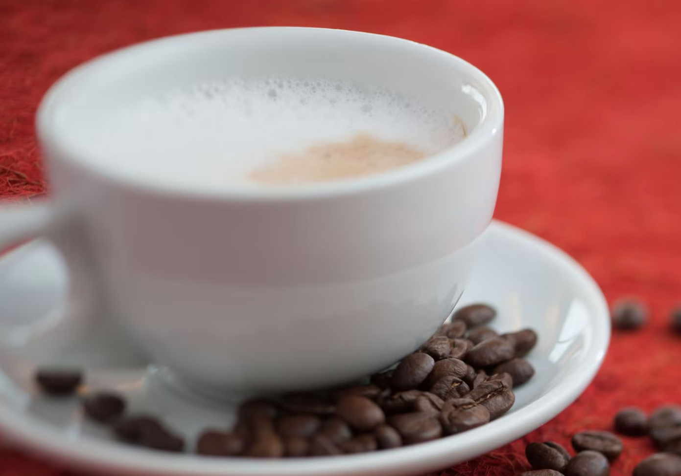 A cup of cappuccino with coffee beans scattered on a white saucer against a red background.