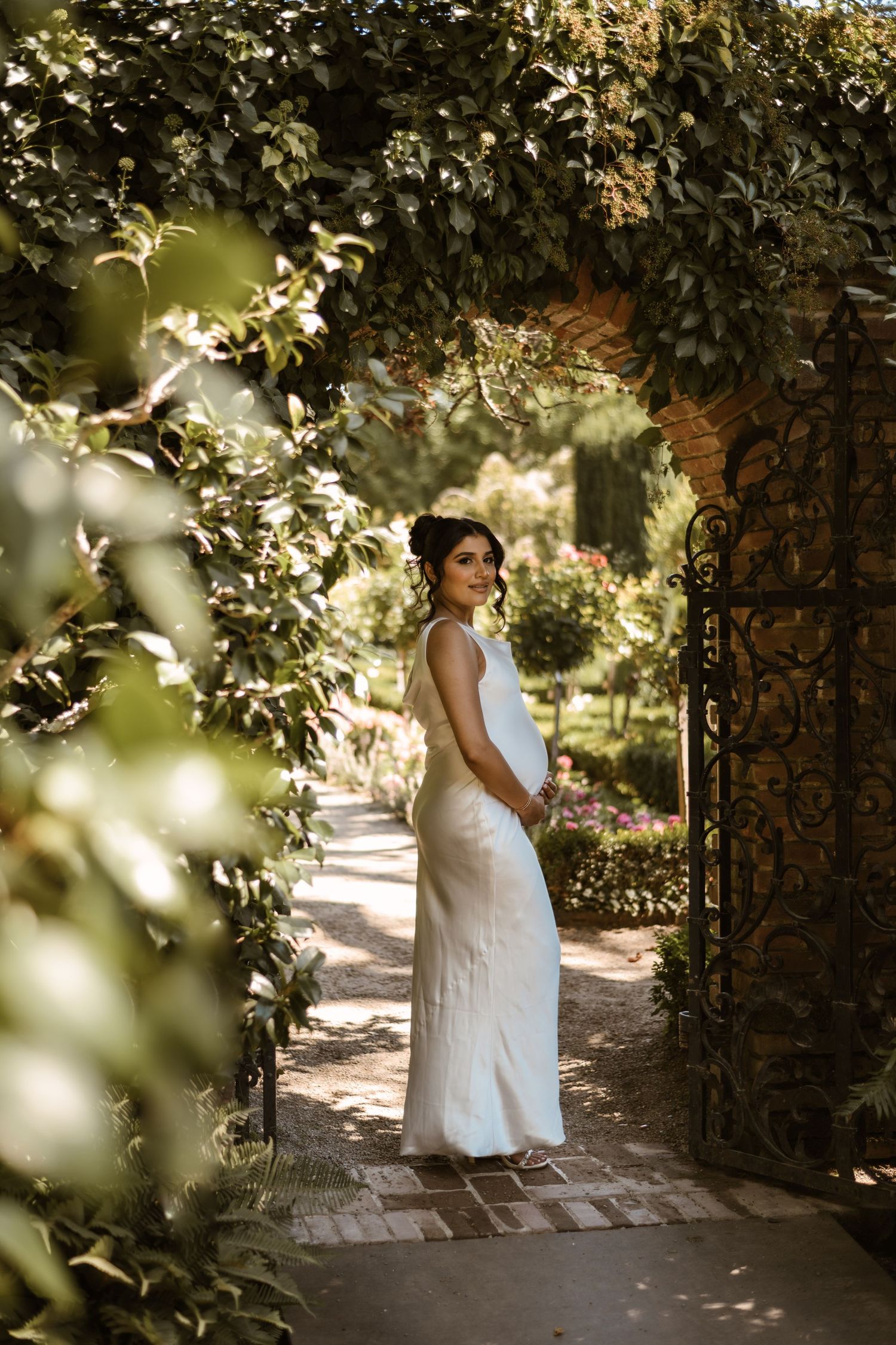 Bride posing in white dress under garden archway with dappled sunlight.