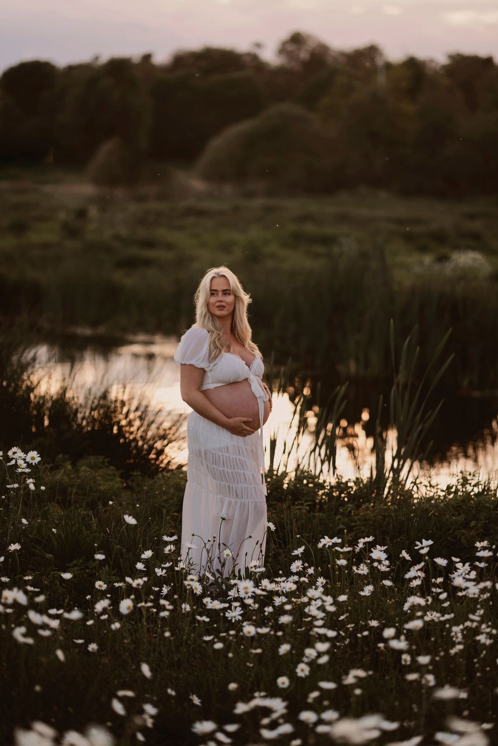 Person in a white dress stands among blooming white daisies near a tranquil pond at sunset.