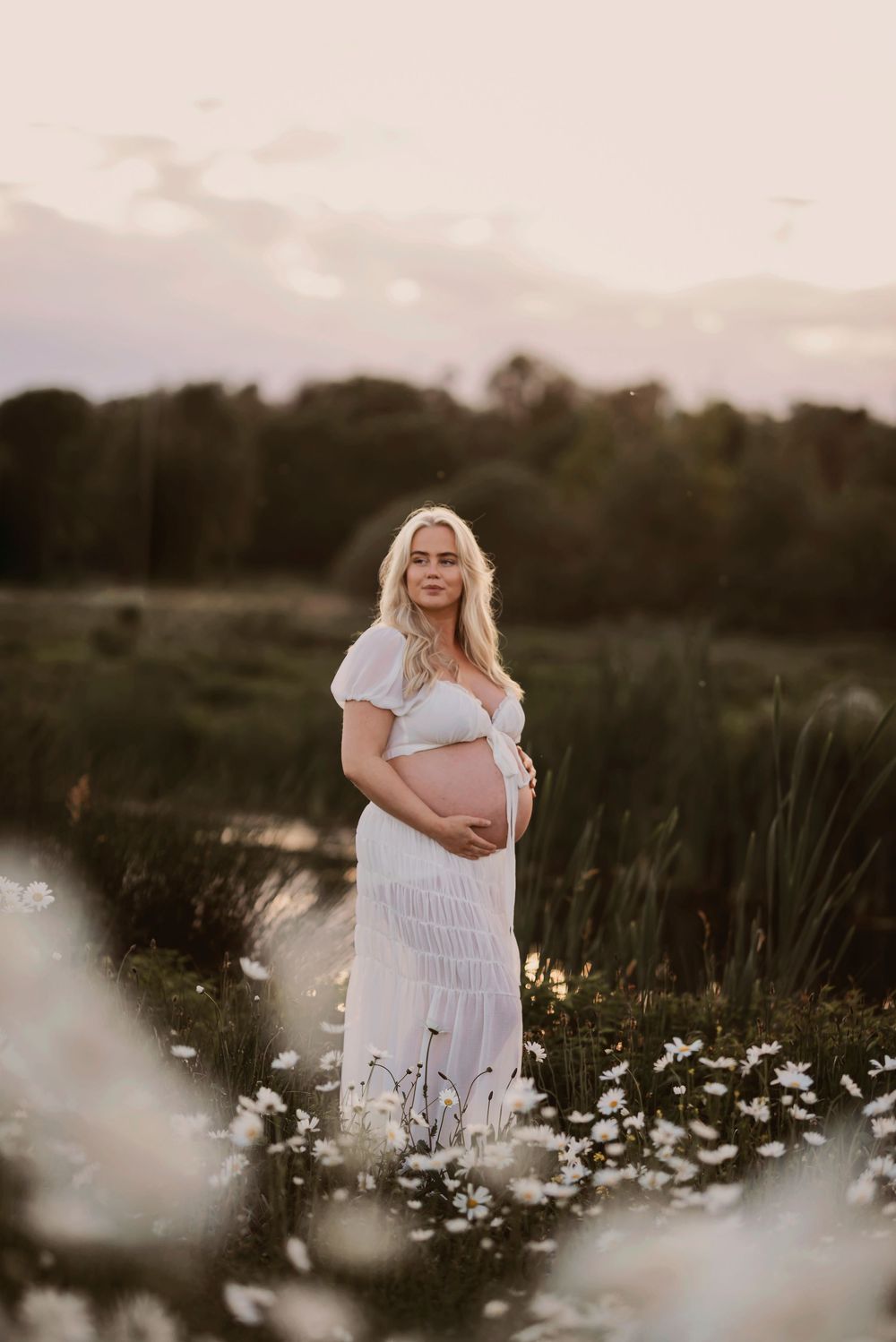 Ethereal maternity photoshoot in a dreamy wildflower meadow at sunset with white daisies and soft lighting.