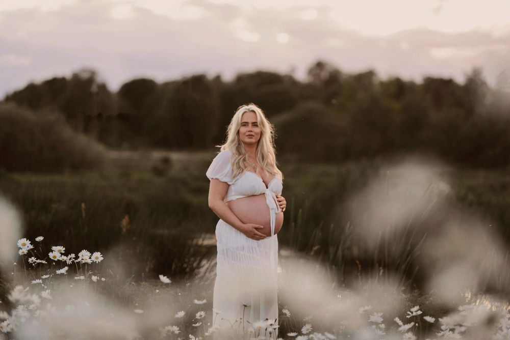 Maternity portrait session in a field of white daisies during golden hour with soft natural lighting.