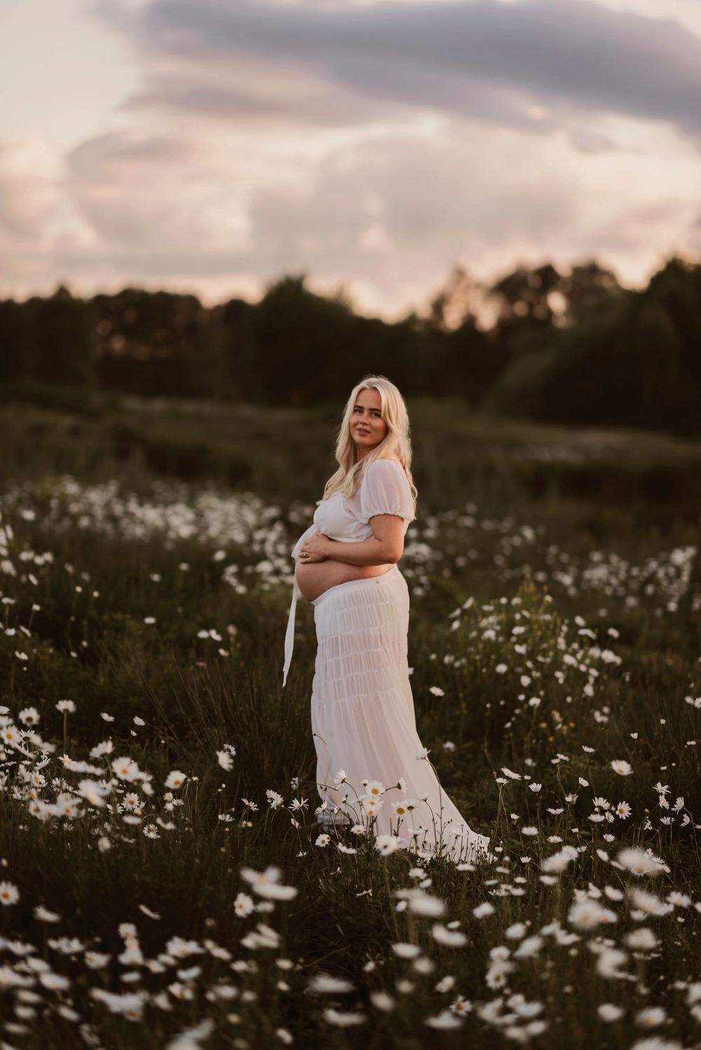 Expectant mother in flowing white dress poses gracefully among blooming daisies at dusk.