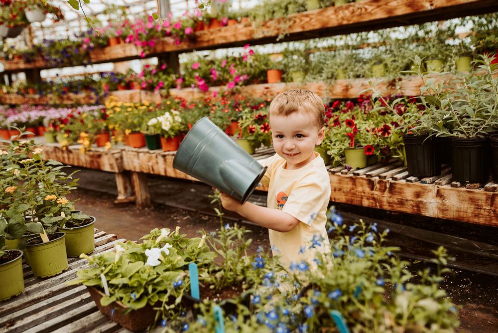 Small helper holding a watering can in a greenhouse filled with potted plants and flowers.