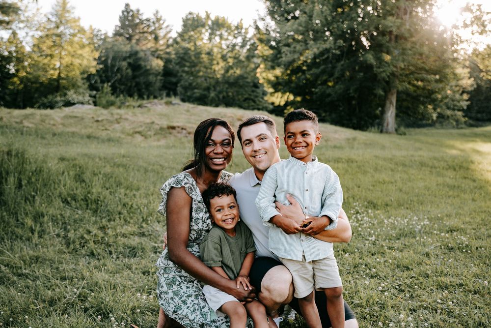 Happy interracial family of four smiling together in a sunny green park, parents with two young boys outdoors.