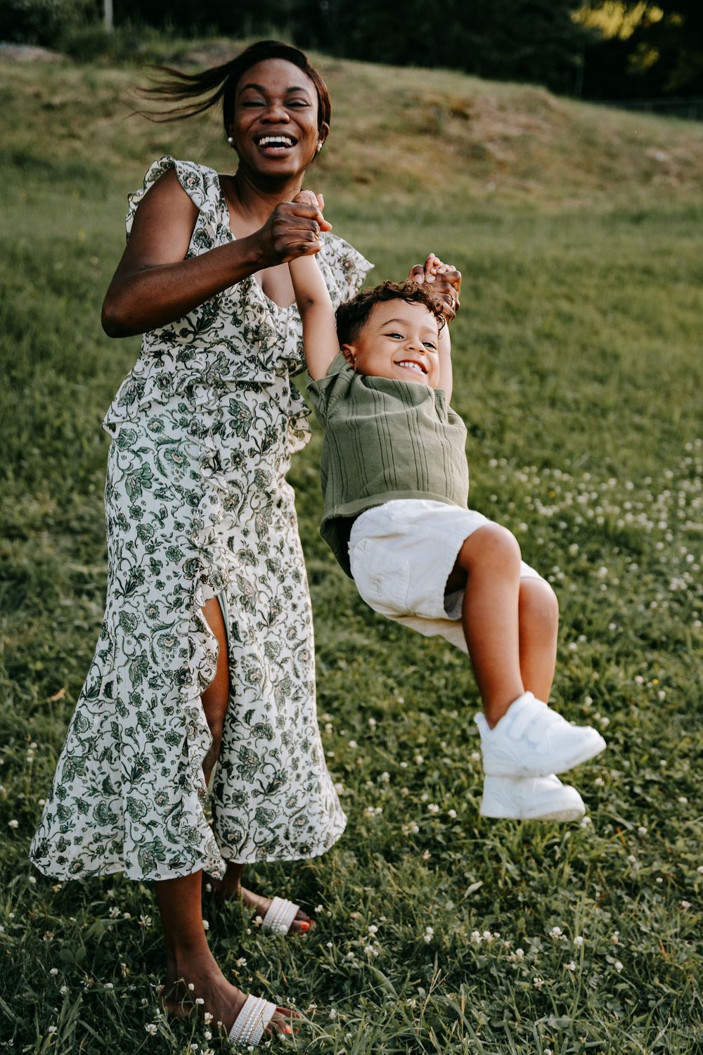 A joyful mother in a floral dress swings her laughing child in a sunny green field.