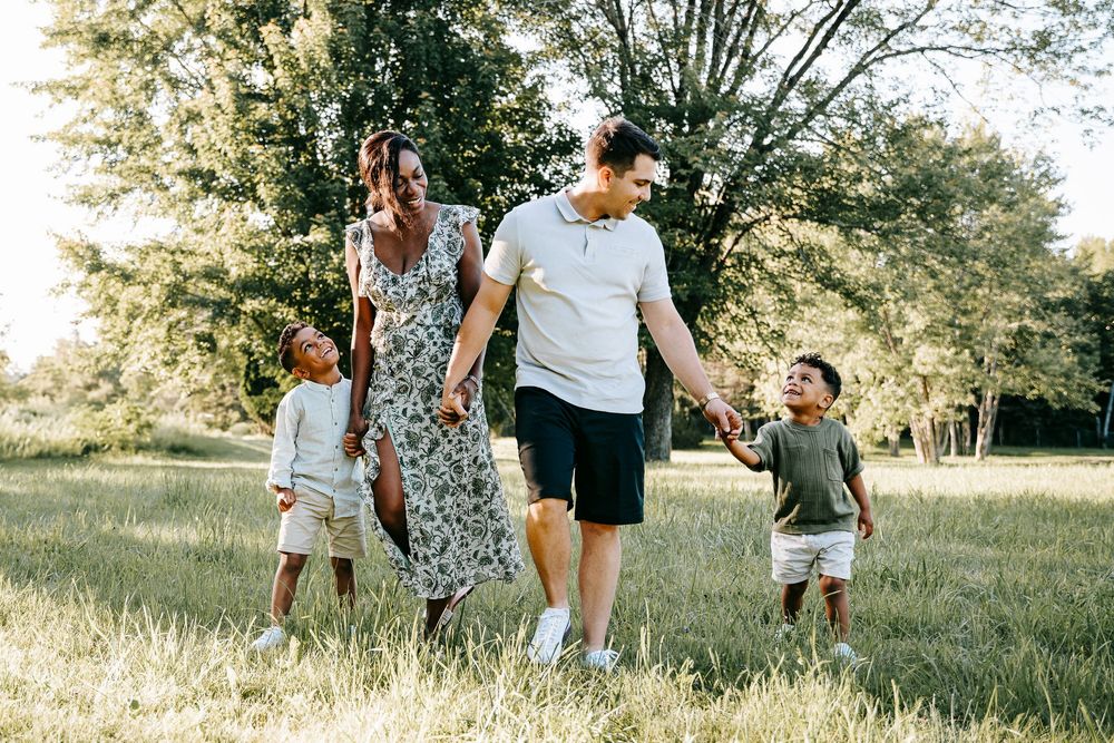 Happy family of four walking in a sunny green park, parents holding hands with two young children.