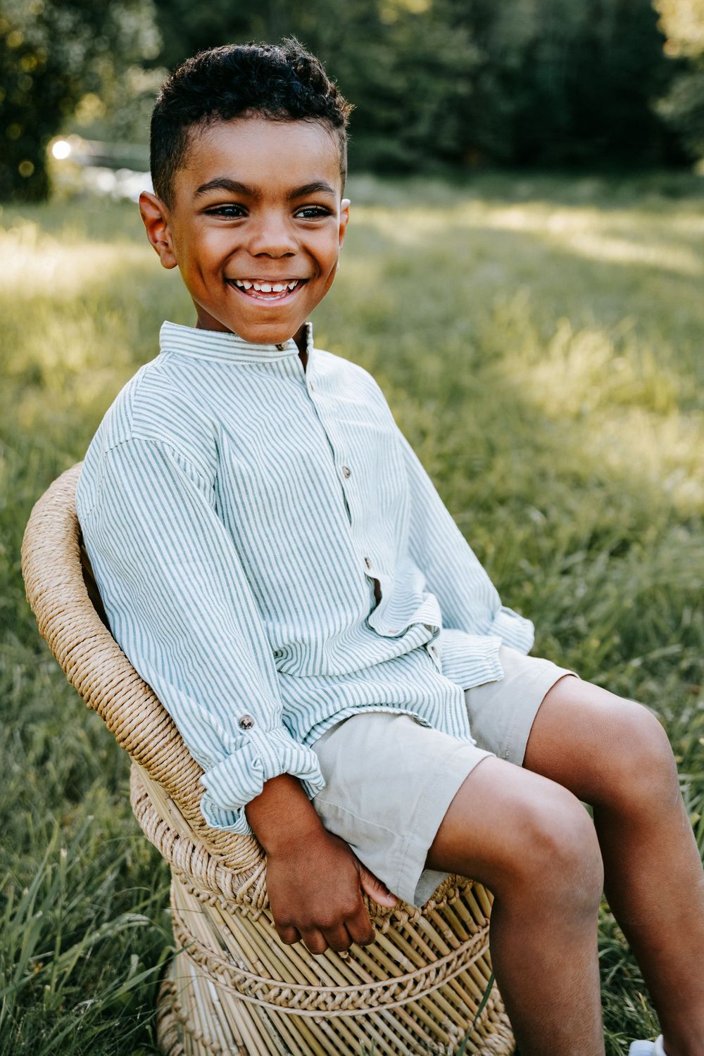 Smiling young boy sitting in a wicker chair outdoors, wearing a striped shirt and khaki shorts in a green field.