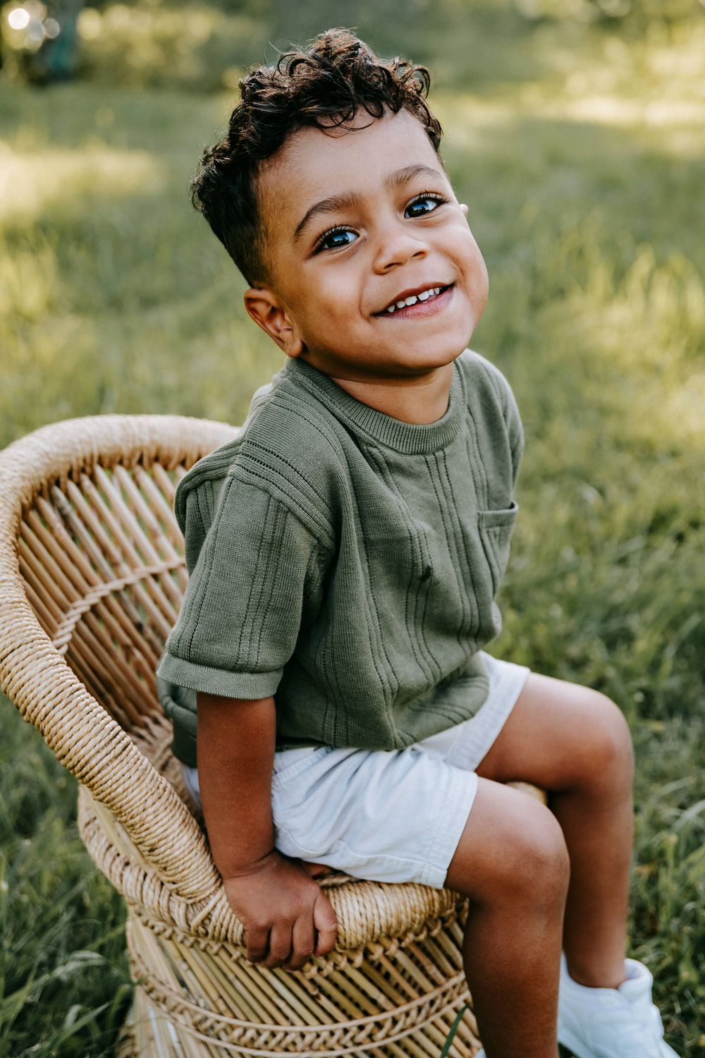 Smiling young boy in green shirt and white shorts sitting on wicker chair outdoors in green grass.