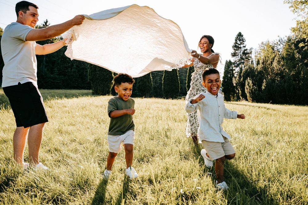 Family playing outdoors in a sunny green field, parents holding a blanket while two children run and laugh together.