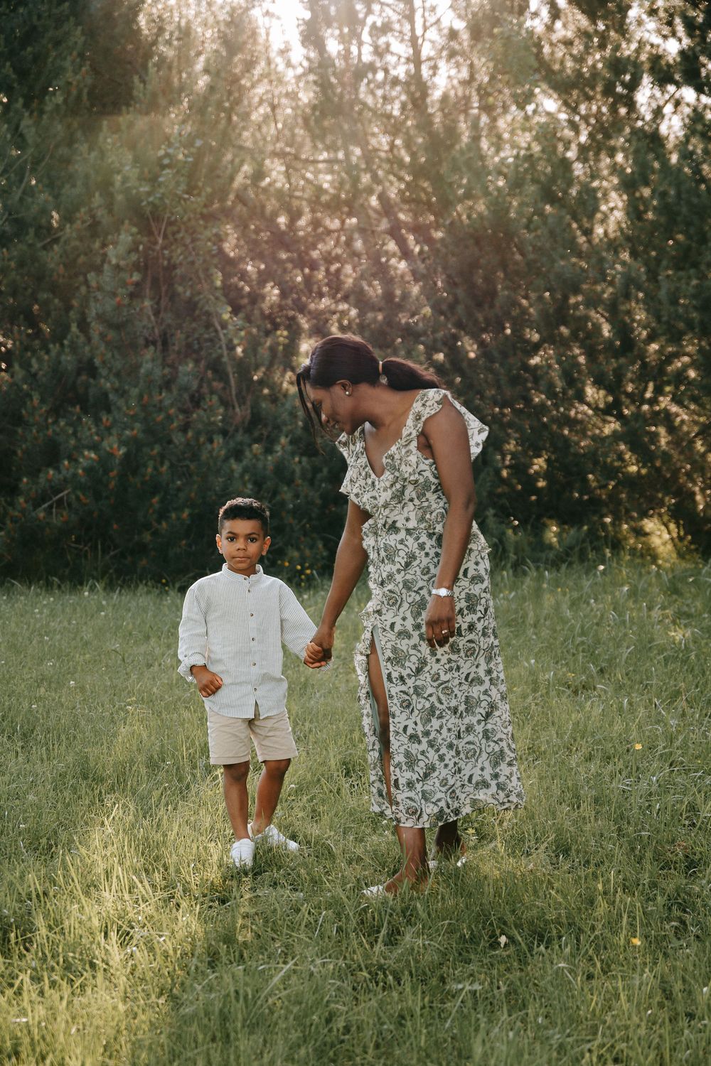 A mother in a floral dress holds hands with her young son in a sunlit green meadow.