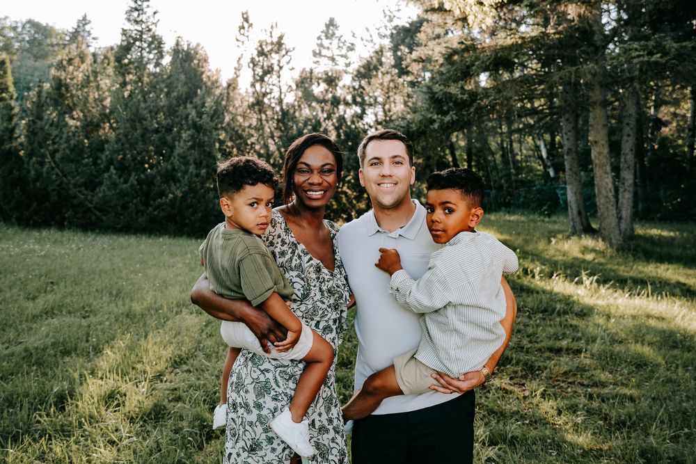 Happy interracial family of four posing outdoors in a green meadow with trees, parents each holding a young child.