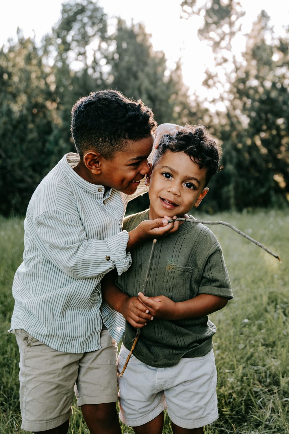 Two young brothers share a joyful moment outdoors, one hugging the other holding a stick.