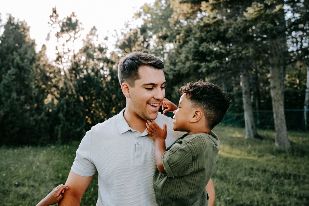 A father smiles warmly while holding his young son outdoors in a lush green park setting.