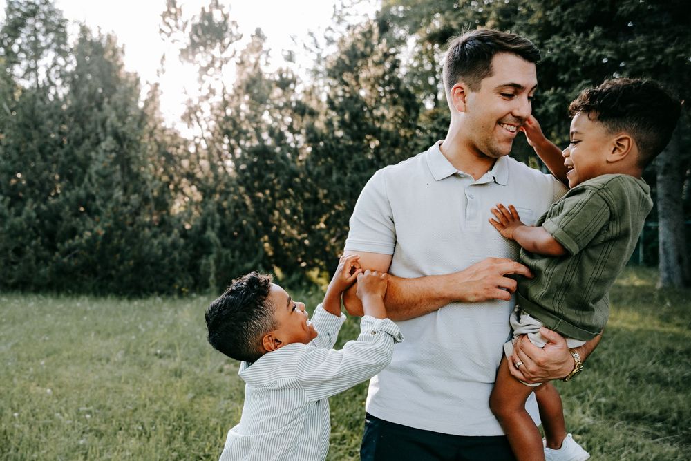 A smiling father holds his toddler son while his older boy plays nearby in a lush green park setting.