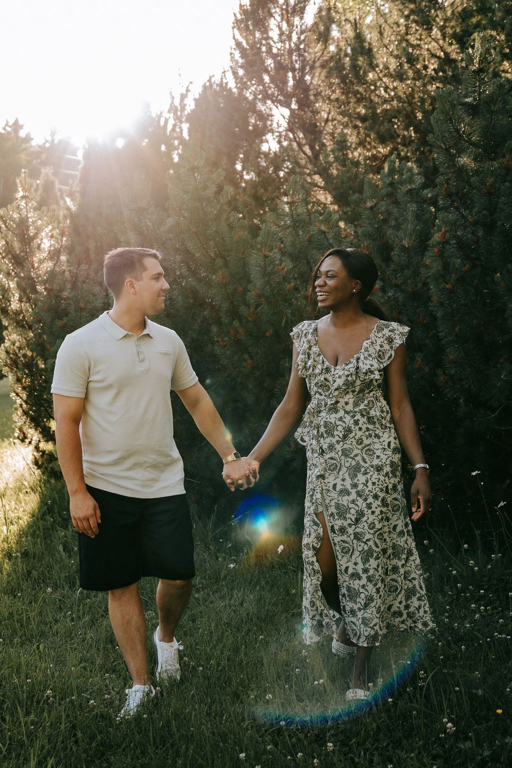A smiling interracial couple holds hands while walking through a sunlit park surrounded by lush green trees.